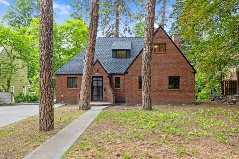 Charming brick Tudor-style home with black windows, nestled among tall trees in a lush green setting.