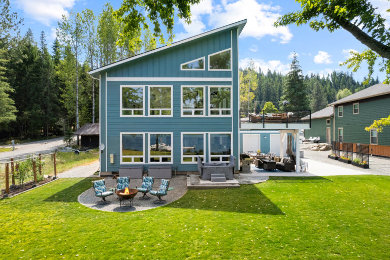 odern blue lakefront home with large windows, outdoor seating, a fire pit, and scenic forest views.